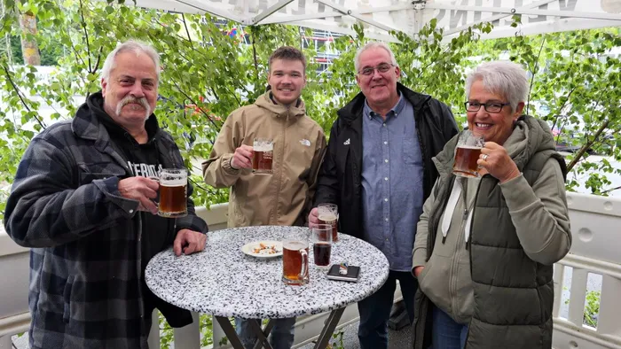 Vier Männer und eine Frau stehen um einen runden Tisch mit Biergläsern im Freien. Sie lächeln in die Kamera und halten ihre Getränke hoch.