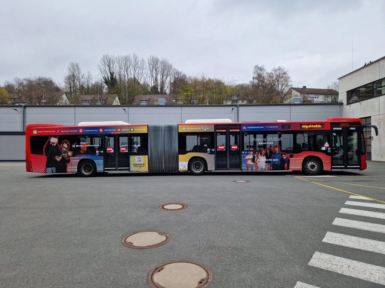Roter Gelenkbus mit der Aufschrift „Tumo“ und Bildern von jungen Menschen, die an Computern arbeiten, steht auf einem Parkplatz vor einem grauen Gebäude.
