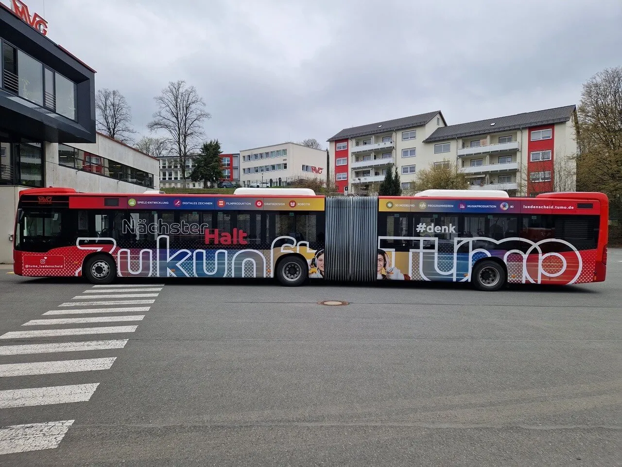 Roter Bus mit der Aufschrift „Zukunft Halt“ und dem Hashtag #denk fährt auf einer Straße vor einem mehrstöckigen Gebäude.