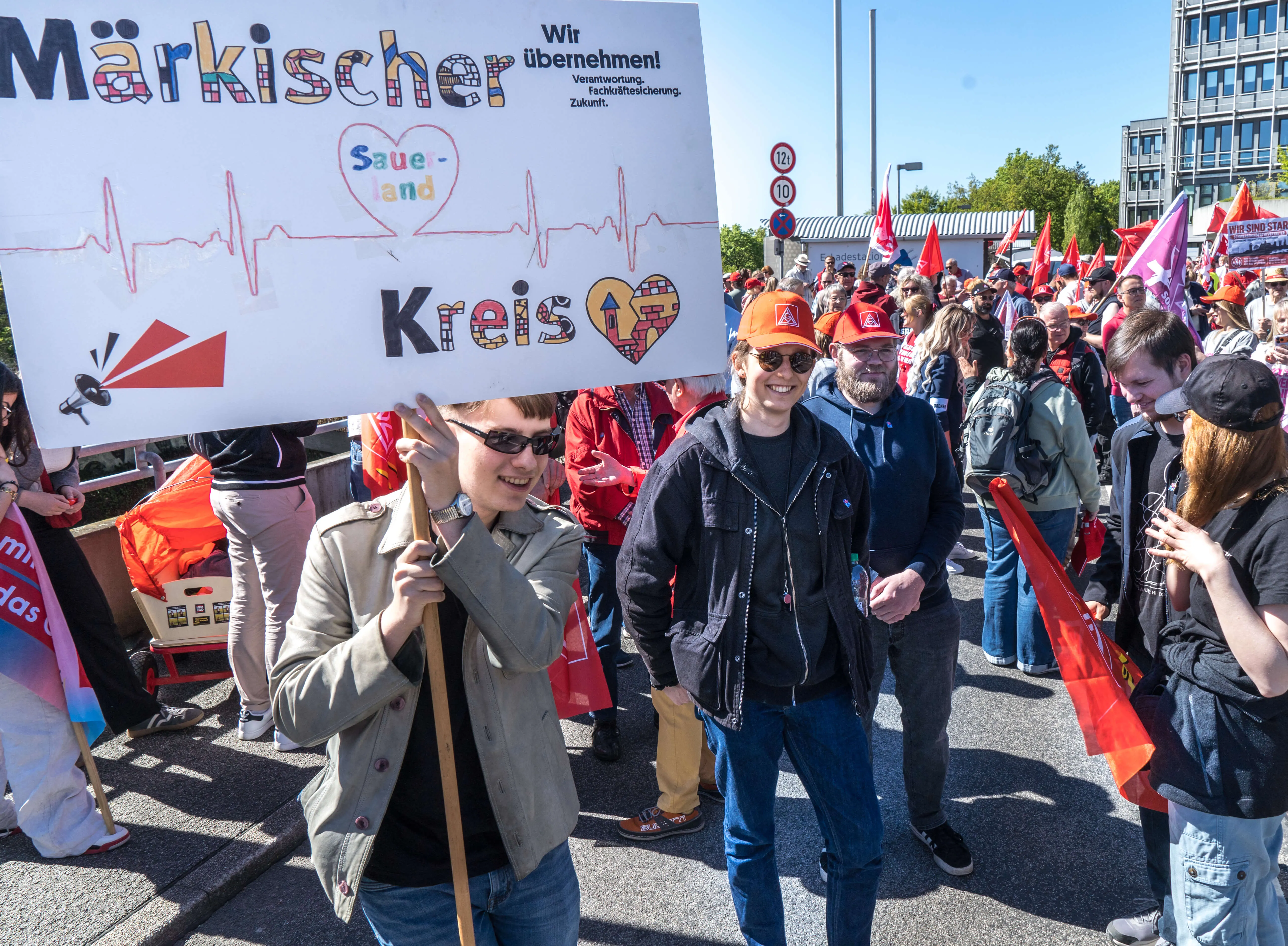 Demonstranten ziehen mit roten Fahnen und Schildern durch eine Stadt, im Vordergrund eine junge Frau mit einem Hund an der Leine und ein lächelnder Mann.
