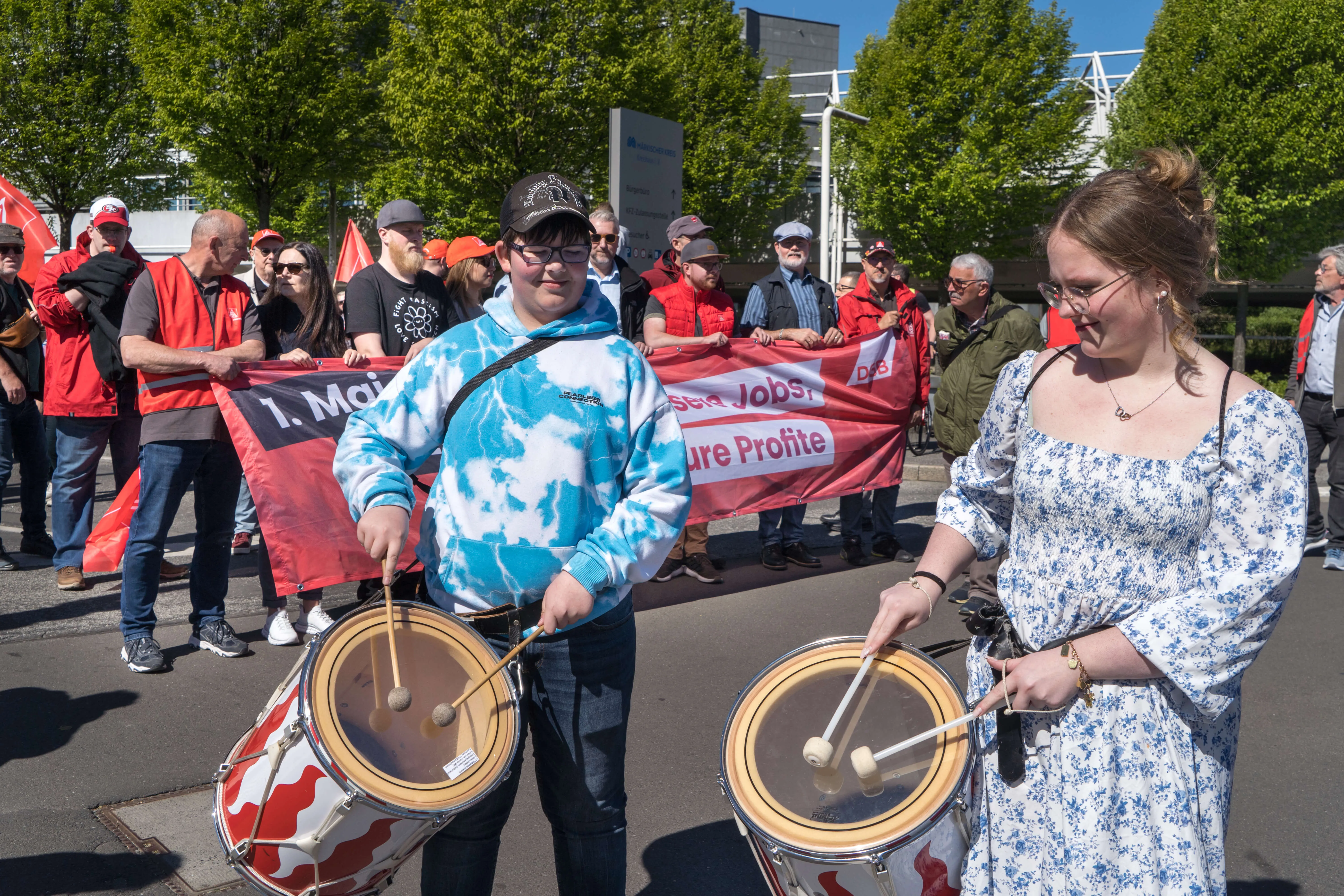Zwei junge Erwachsene spielen Trommeln an der Spitze einer Demonstration mit roten Fahnen und Transparenten mit der Aufschrift „1. Mai“ und „Jobs sichern, faire Profite“.