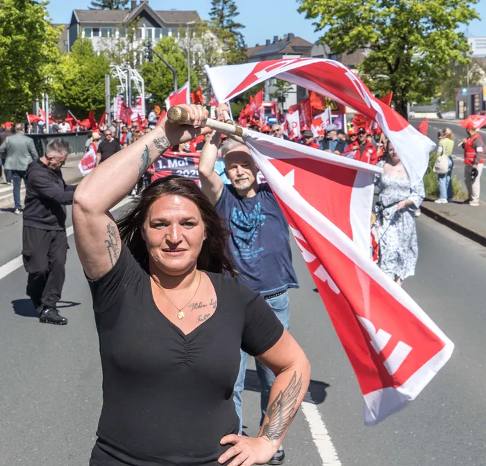 Eine Frau mit Tätowierungen auf den Armen hält eine rote Fahne über ihren Kopf, während sie an einer Demonstration teilnimmt. Im Hintergrund sind weitere Demonstrierende und rote Fahnen zu sehen.