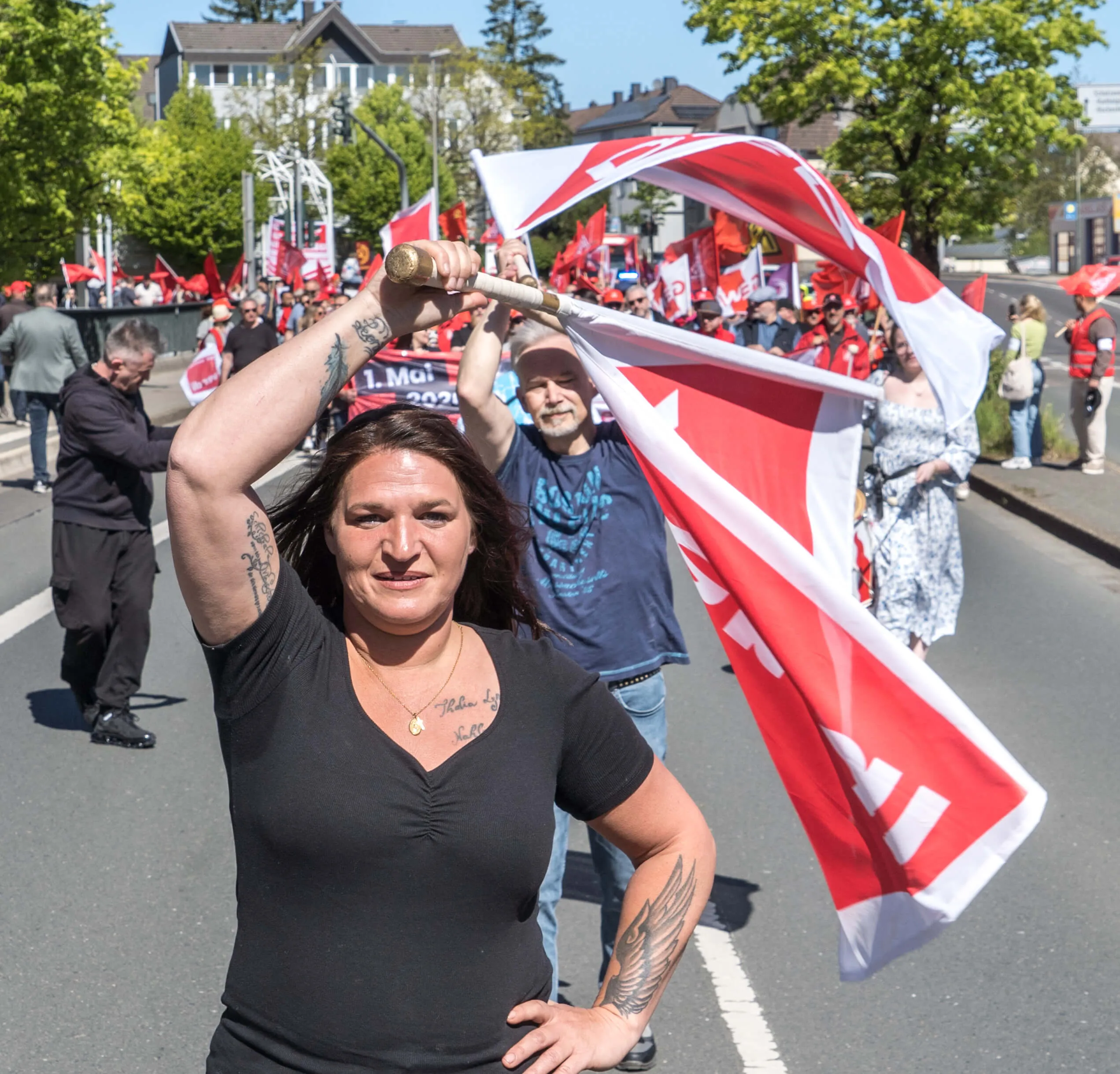 Eine Frau mit Tätowierungen auf den Armen hält eine rote Fahne über ihren Kopf, während sie an einer Demonstration teilnimmt. Im Hintergrund sind weitere Demonstrierende und rote Fahnen zu sehen.
