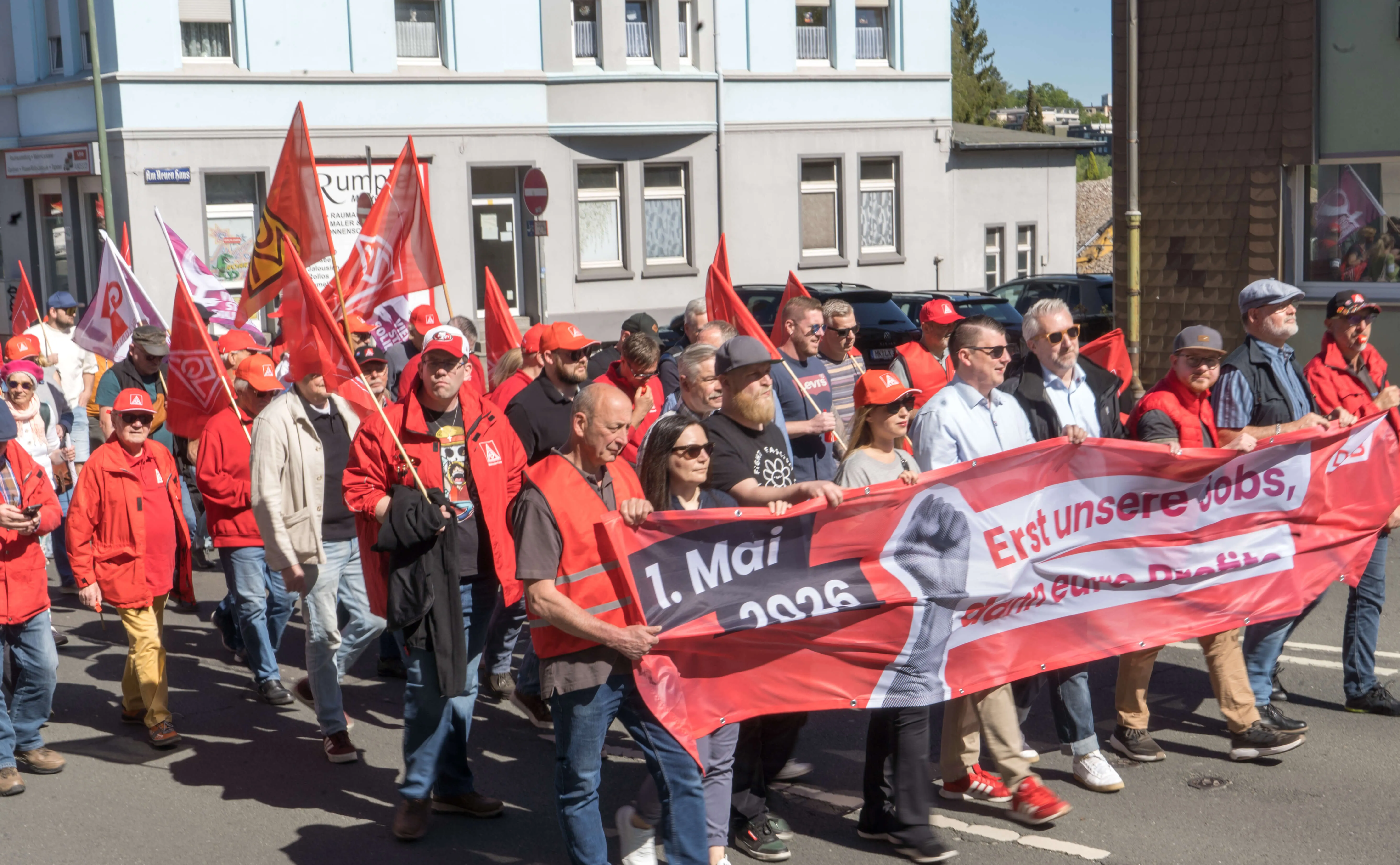 Demonstrationszug mit vielen Menschen, die rote Fahnen und ein Banner mit der Aufschrift „1. Mai 2026 Erst unsere Jobs“ tragen.