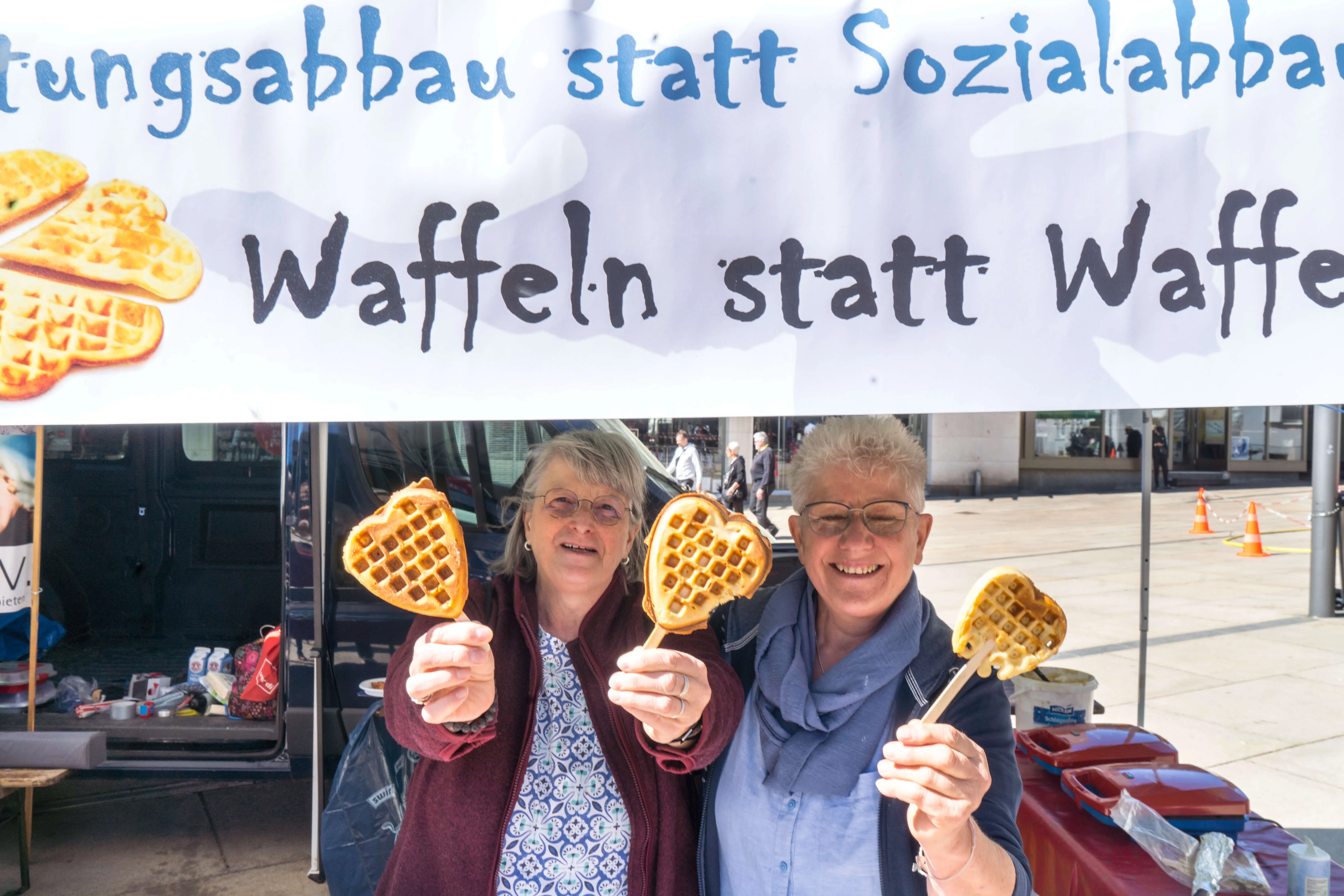 Zwei Frauen halten Waffeln vor sich hoch, im Hintergrund ein Banner mit der Aufschrift „Waffeln statt Waffe“.