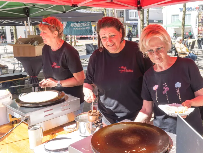 Drei Frauen bereiten auf einem Stand Crêpes zu, während im Hintergrund eine Menschenmenge auf dem Rathausplatz zu sehen ist.
