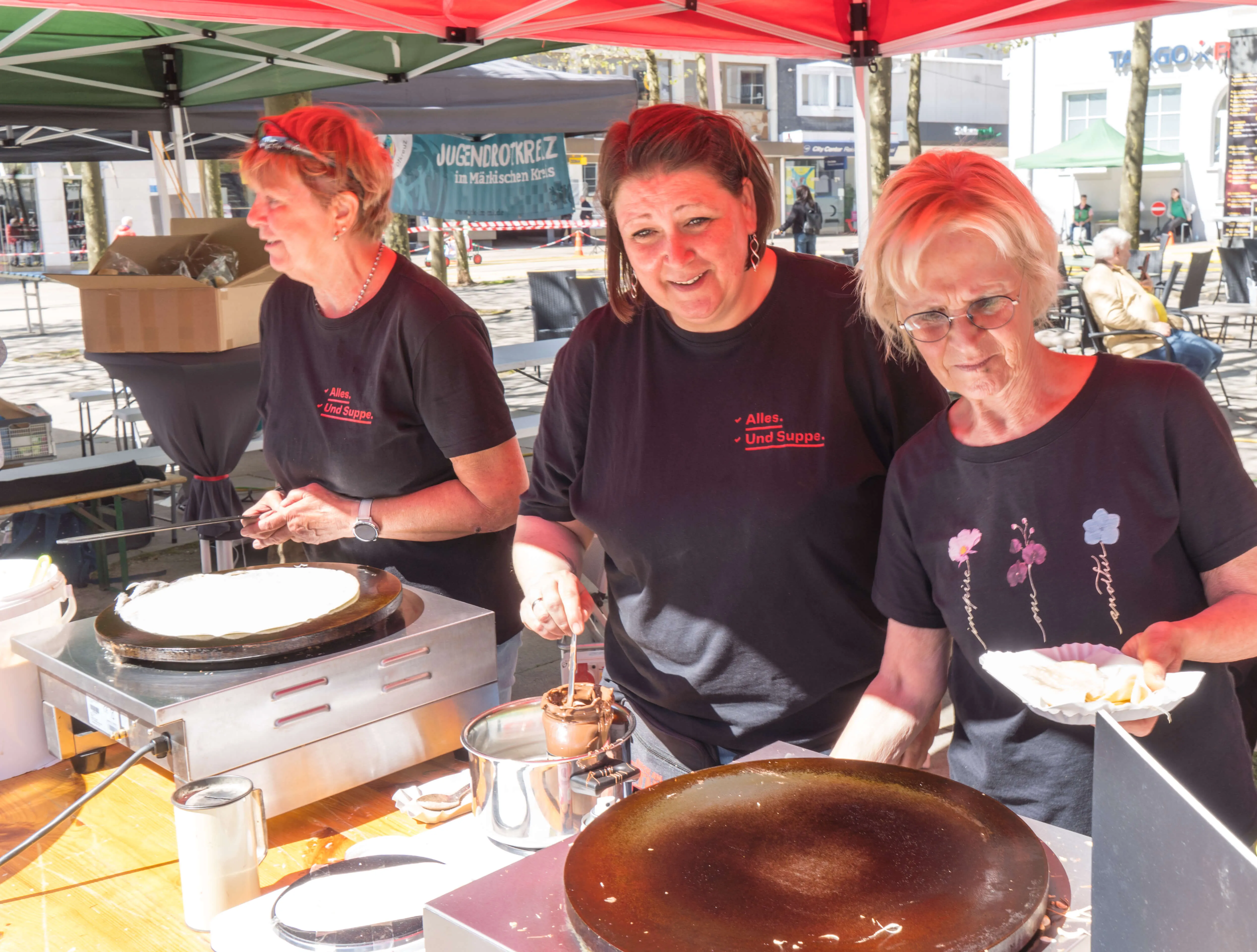 Drei Frauen bereiten auf einem Stand Crêpes zu, während im Hintergrund eine Menschenmenge auf dem Rathausplatz zu sehen ist.