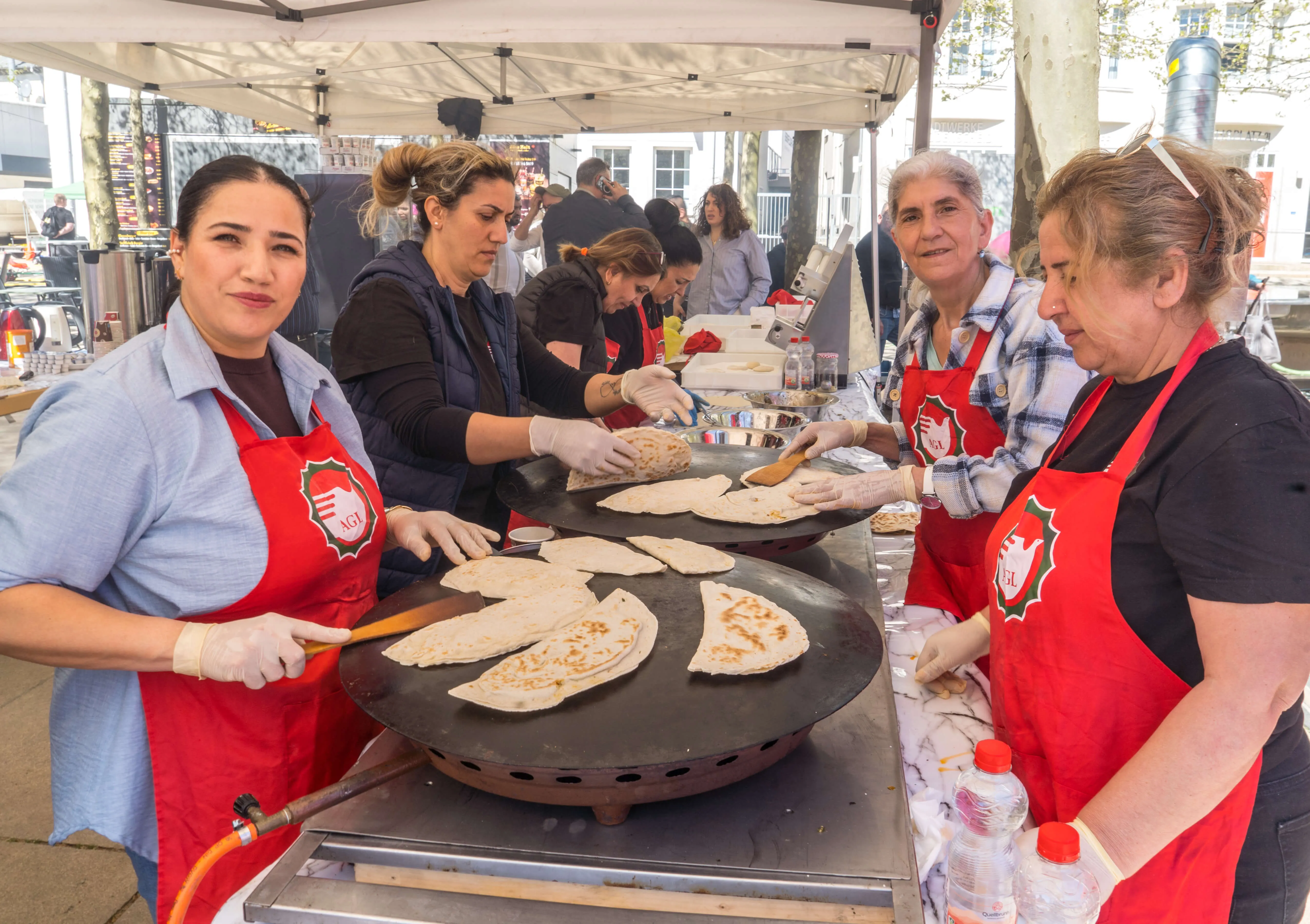 Mehrere Frauen in roten Schürzen bereiten auf einem großen Grill Fladenbrot zu, während im Hintergrund weitere Menschen an einem Stand stehen.