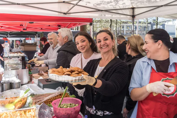 Eine Frau reicht lächelnd eine Platte mit Gebäck über einen Verkaufstresen an eine andere Person. Im Hintergrund stehen weitere Menschen auf dem belebten Rathausplatz.