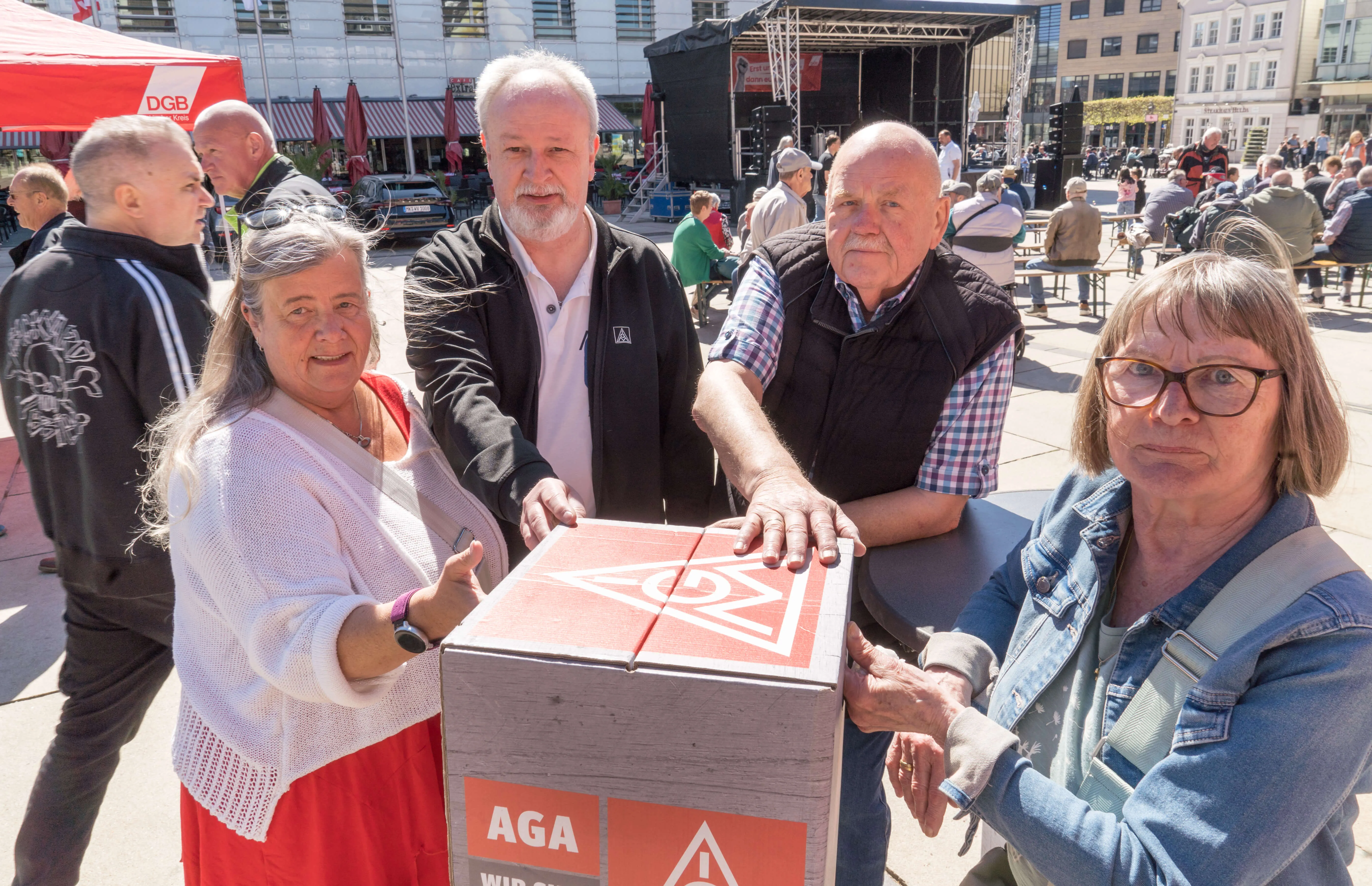 Vier Menschen tragen gemeinsam eine große Wahlurne mit dem Logo einer Partei auf dem Lüdenscheider Rathausplatz. Im Hintergrund sind weitere Kundgebungsteilnehmer und eine Bühne zu sehen.