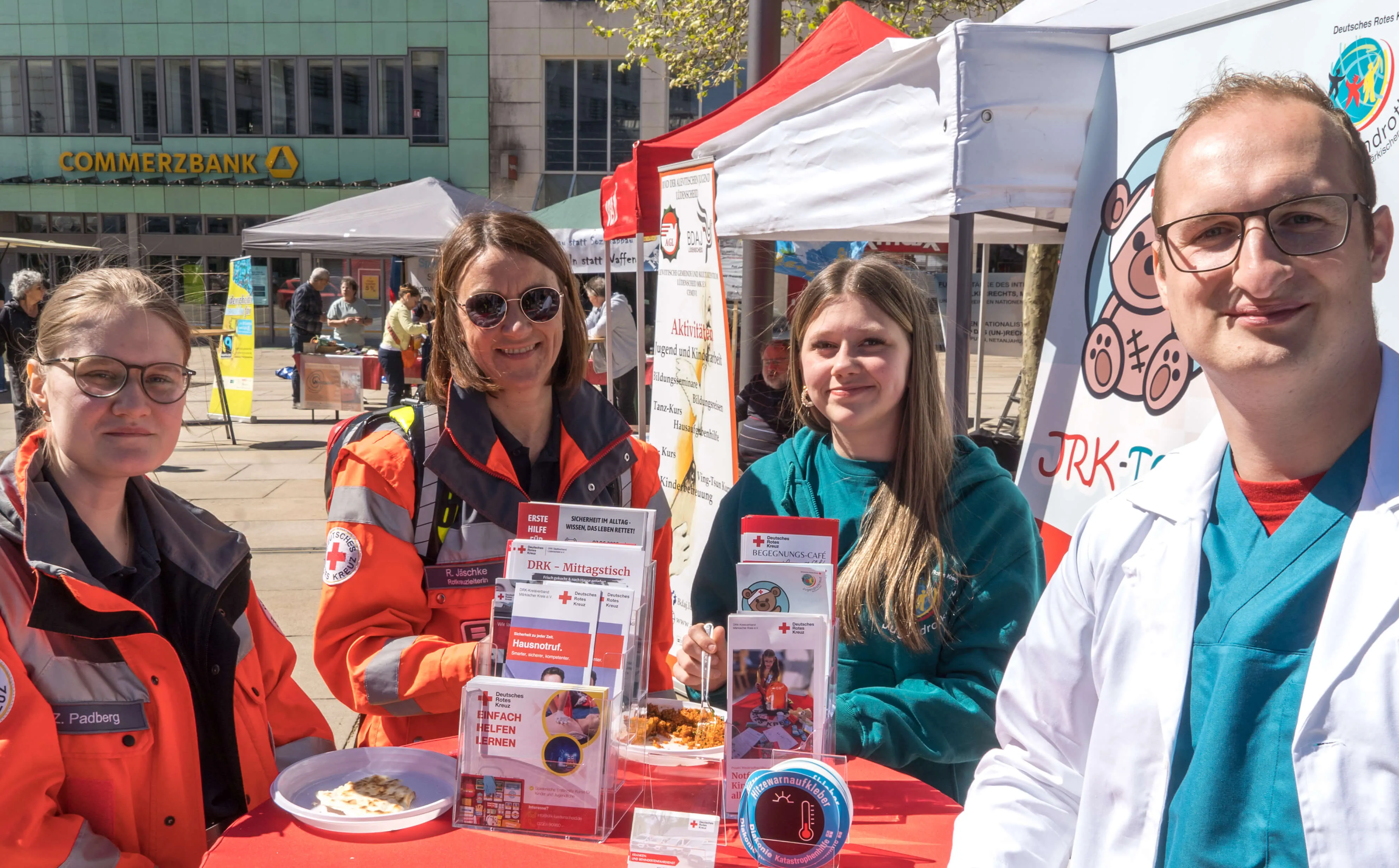Vier ehrenamtliche Helfer des Deutschen Roten Kreuzes stehen an einem Infostand mit Speisen und Informationsmaterialien auf dem Lüdenscheider Rathausplatz.