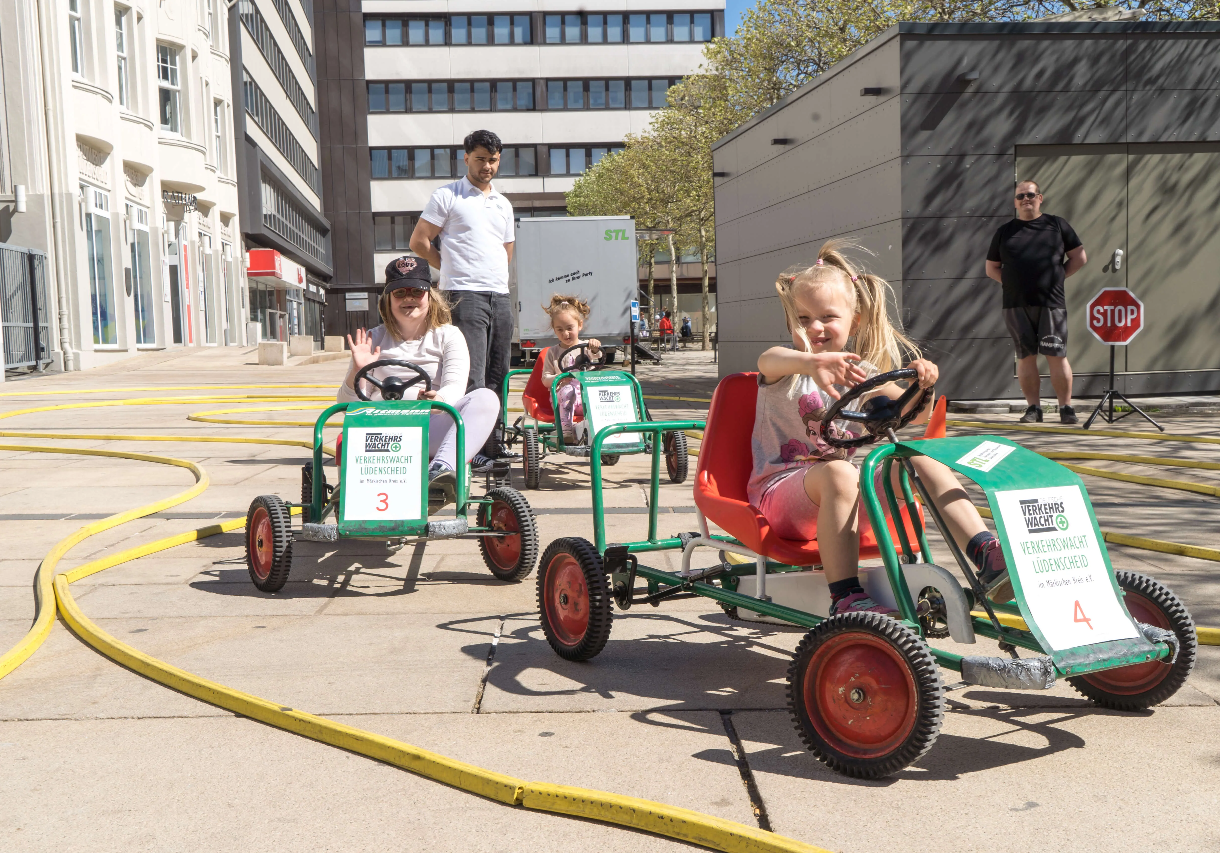 Kinder fahren auf selbstgebauten Karts auf dem Lüdenscheider Rathausplatz, während ein Mann im Hintergrund zuschaut. Die Karts tragen die Nummern 3 und 4.