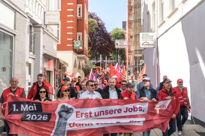 Demonstrationszug mit vielen Menschen, die ein Banner mit der Aufschrift „1. Mai 2026. Erst unsere Jobs“ tragen.
