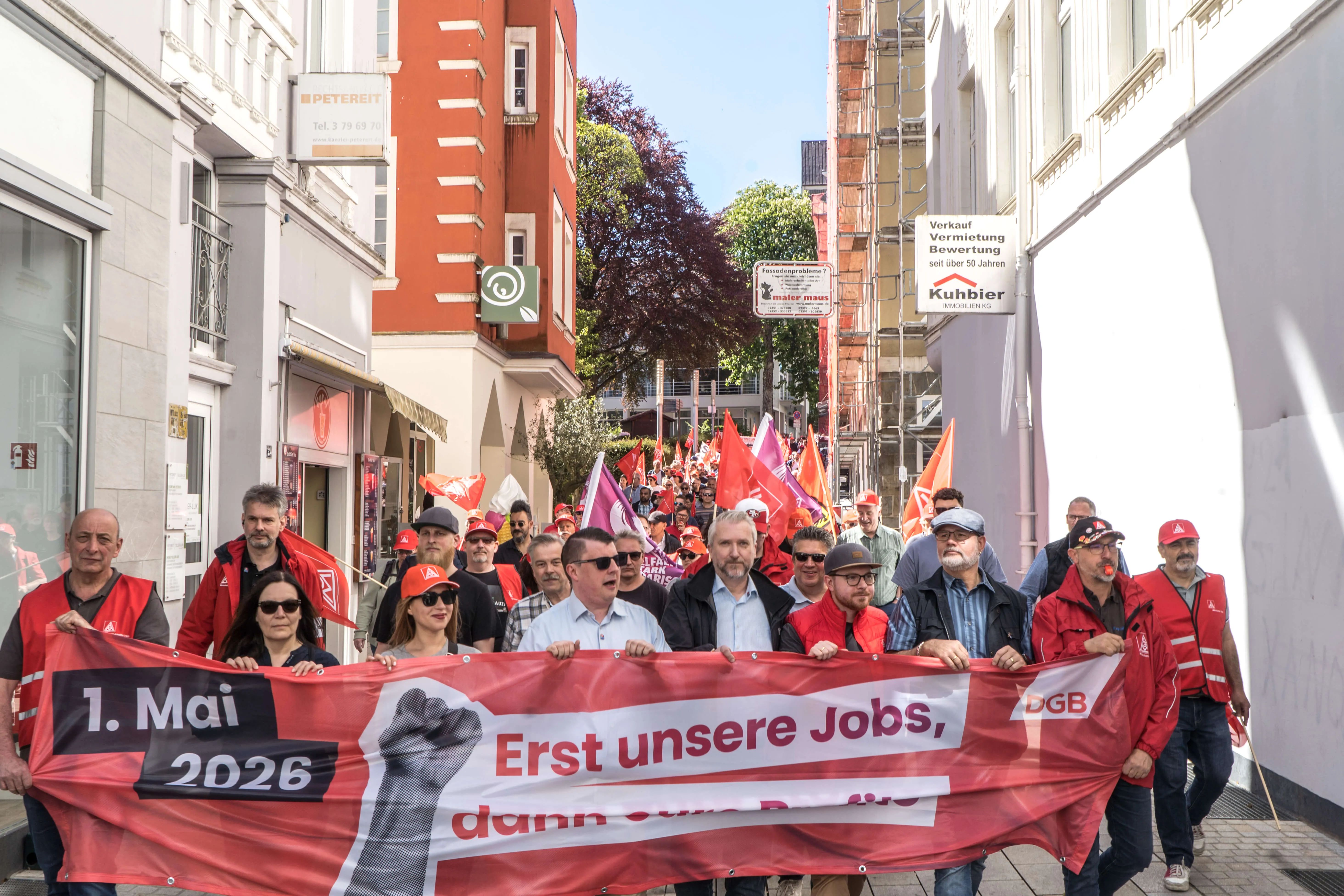 Demonstrationszug mit vielen Menschen, die ein Banner mit der Aufschrift „1. Mai 2026. Erst unsere Jobs“ tragen.
