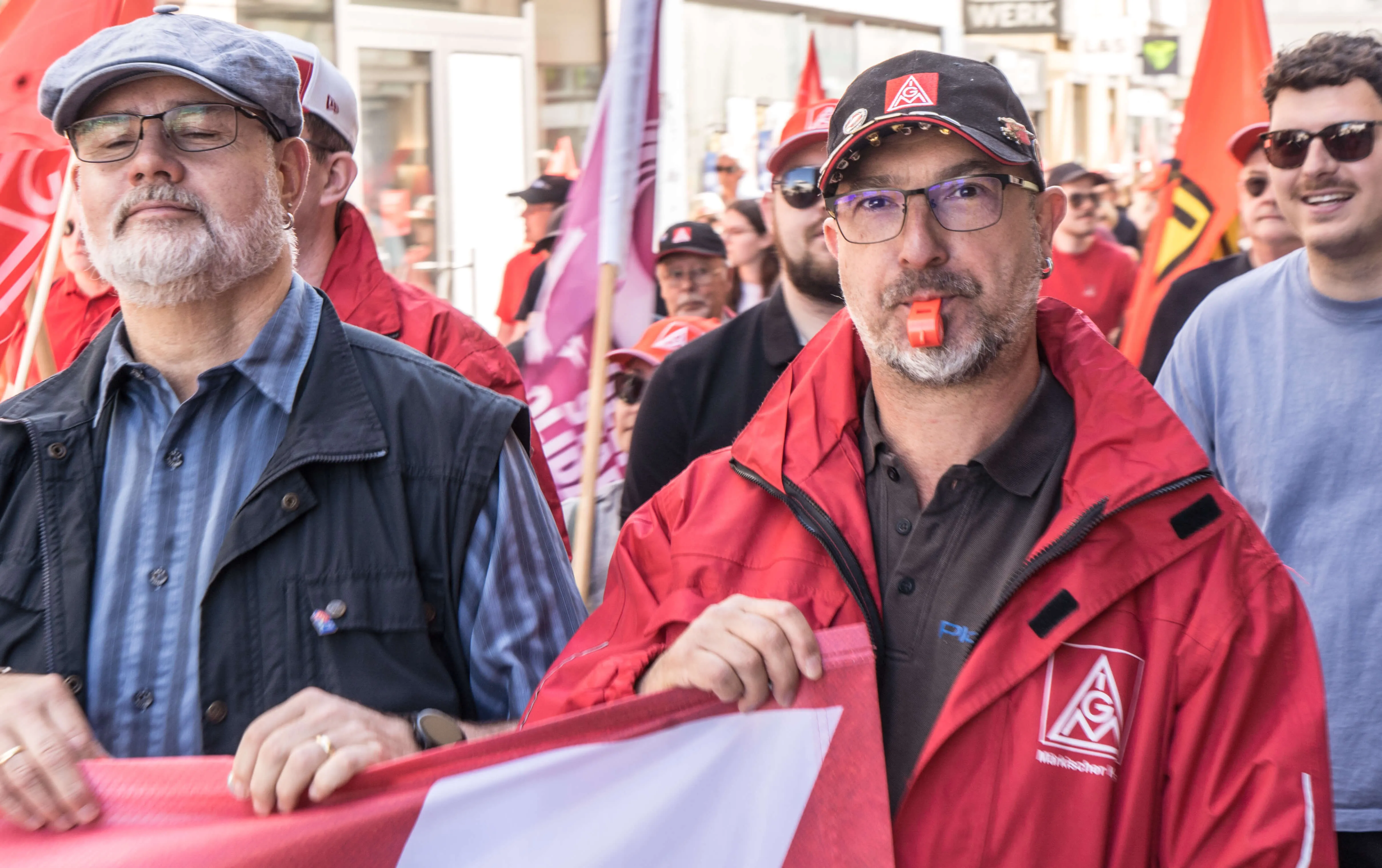 Zwei Männer im Vordergrund, einer mit Bart und Kappe, der andere mit roter Jacke und Kappe, halten während einer Demonstration auf dem Rathausplatz Fahnen. Im Hintergrund weitere Demonstrierende.