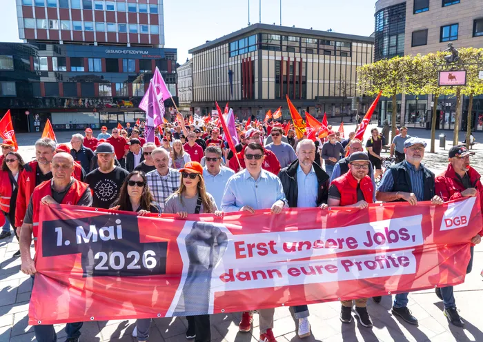 Demonstranten ziehen mit einem roten Banner durch die Menge, auf dem steht „1. Mai 2026 Erst unsere Jobs, dann eure Profite“.