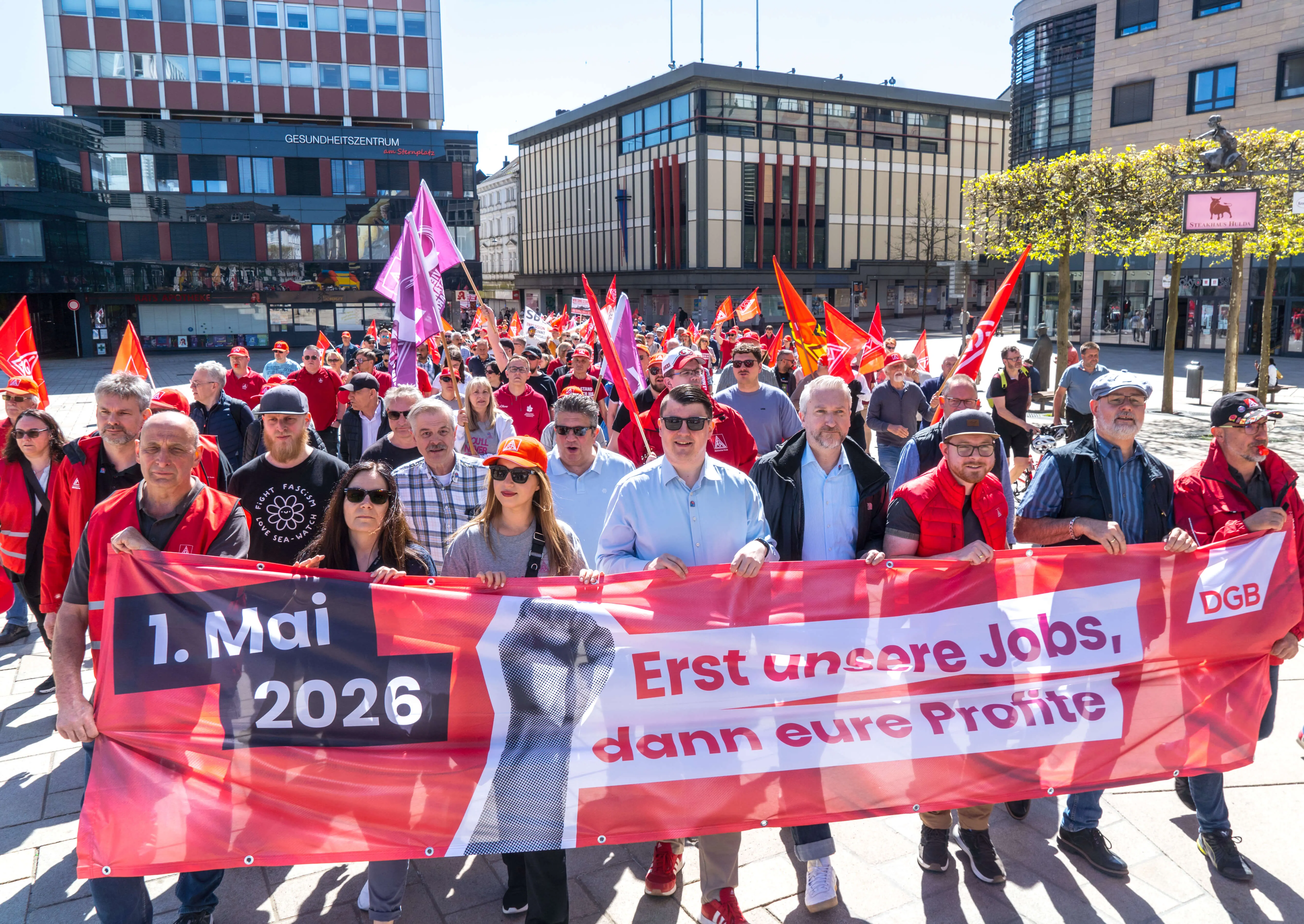 Demonstranten ziehen mit einem roten Banner durch die Menge, auf dem steht „1. Mai 2026 Erst unsere Jobs, dann eure Profite“.