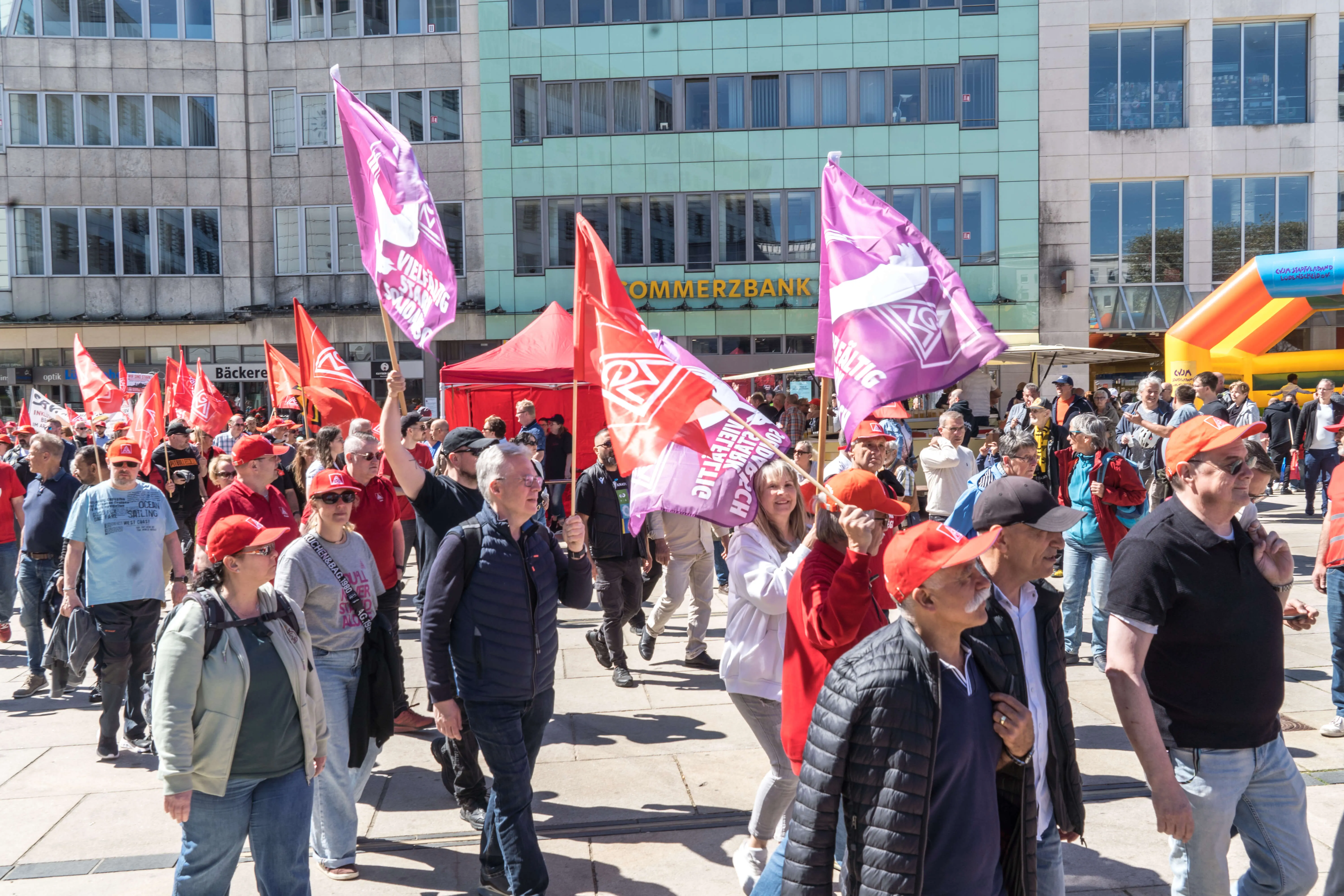 Menschen ziehen mit rot-weiß gefärbten Fahnen und Transparenten über einen Platz vor einem Hochhaus. Viele Teilnehmer tragen rote Kopfbedeckungen.