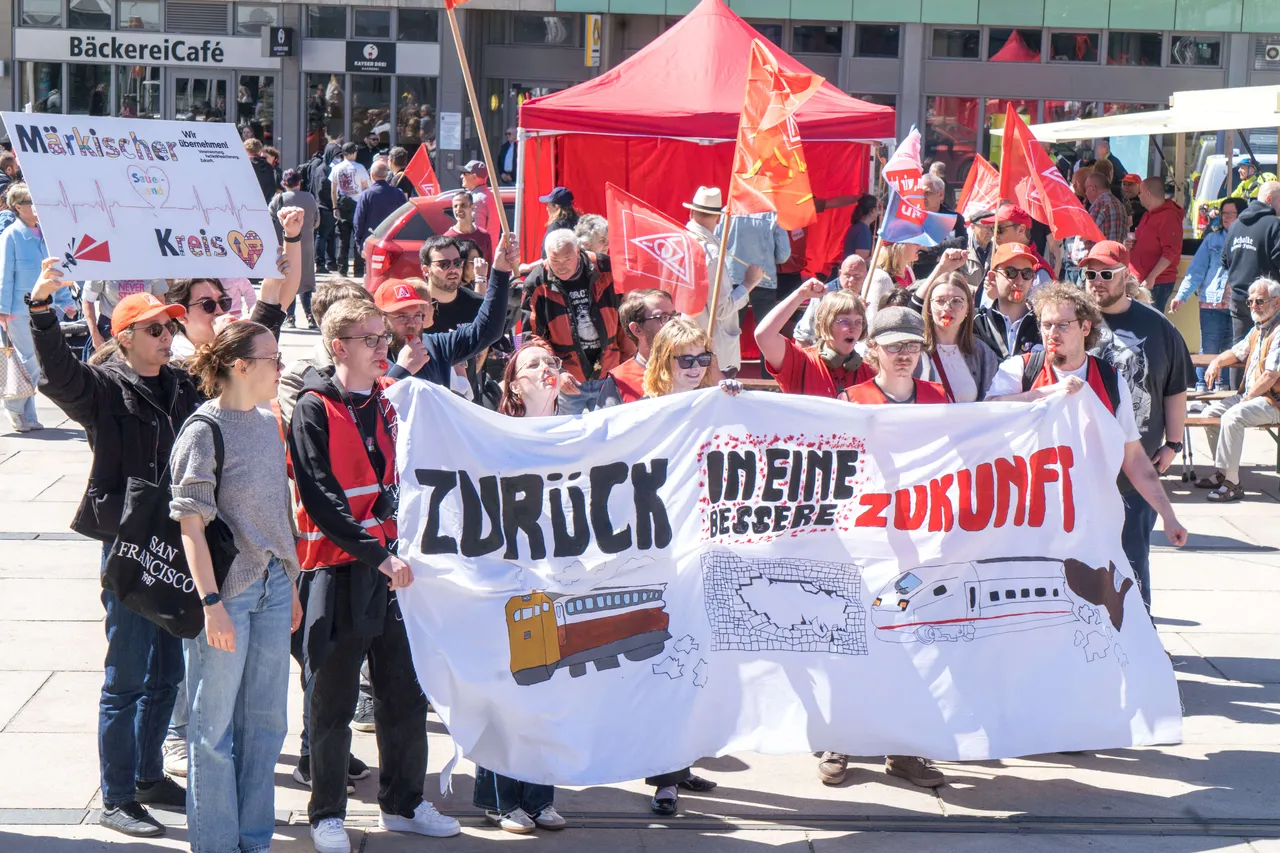Demonstranten tragen ein Banner mit der Aufschrift „Zurück in meine bessere Zukunft“ und Abbildungen eines Schulbusses und eines Krankenwagens. Im Hintergrund ein rotes Zelt und weitere Demonstranten mit roten Fahnen.