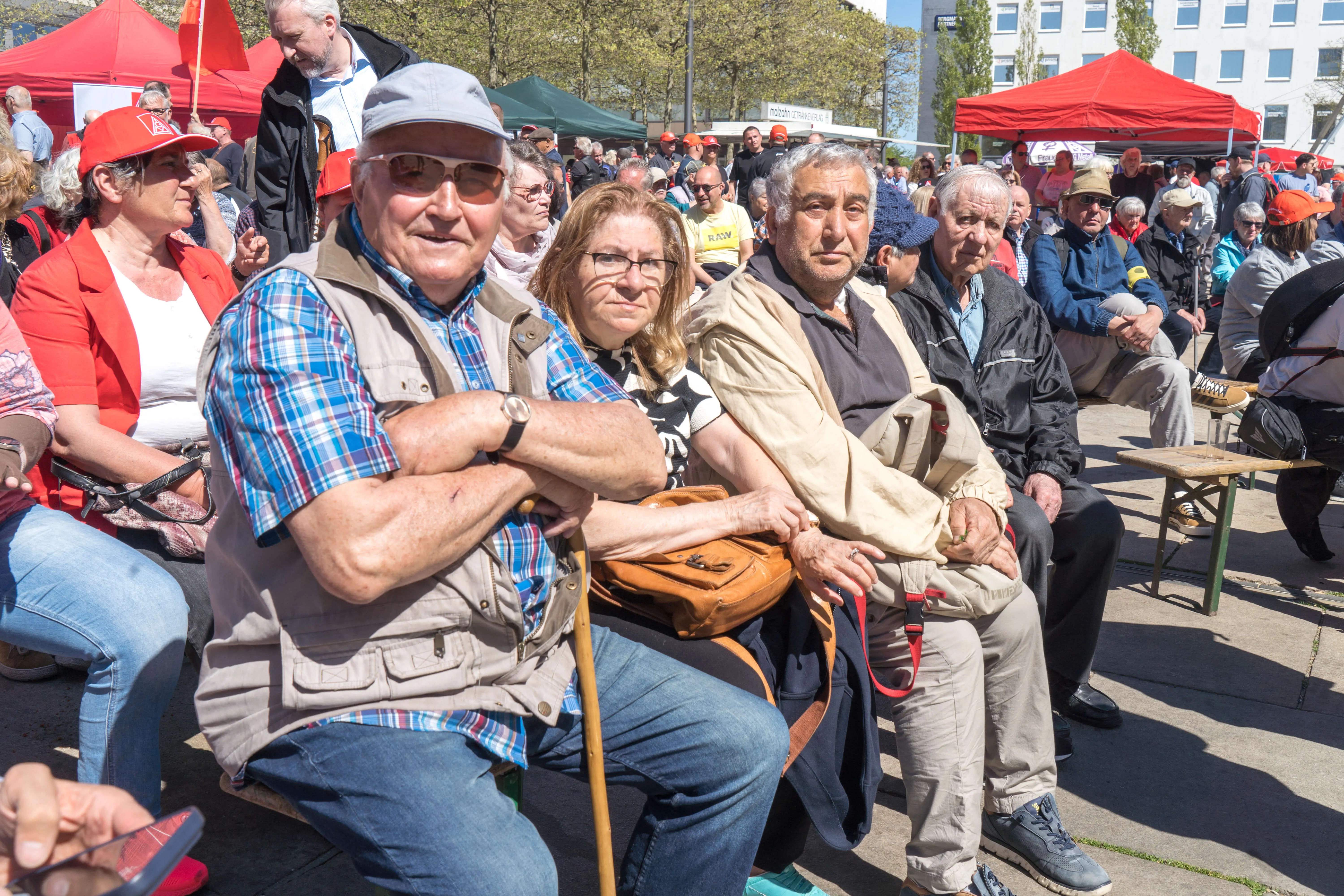 Mehrere ältere Menschen sitzen auf einer Bank und schauen auf eine Menschenmenge, die sich vor einem Rathaus versammelt hat. Im Hintergrund sind rote Zelte und Fahnen zu sehen.