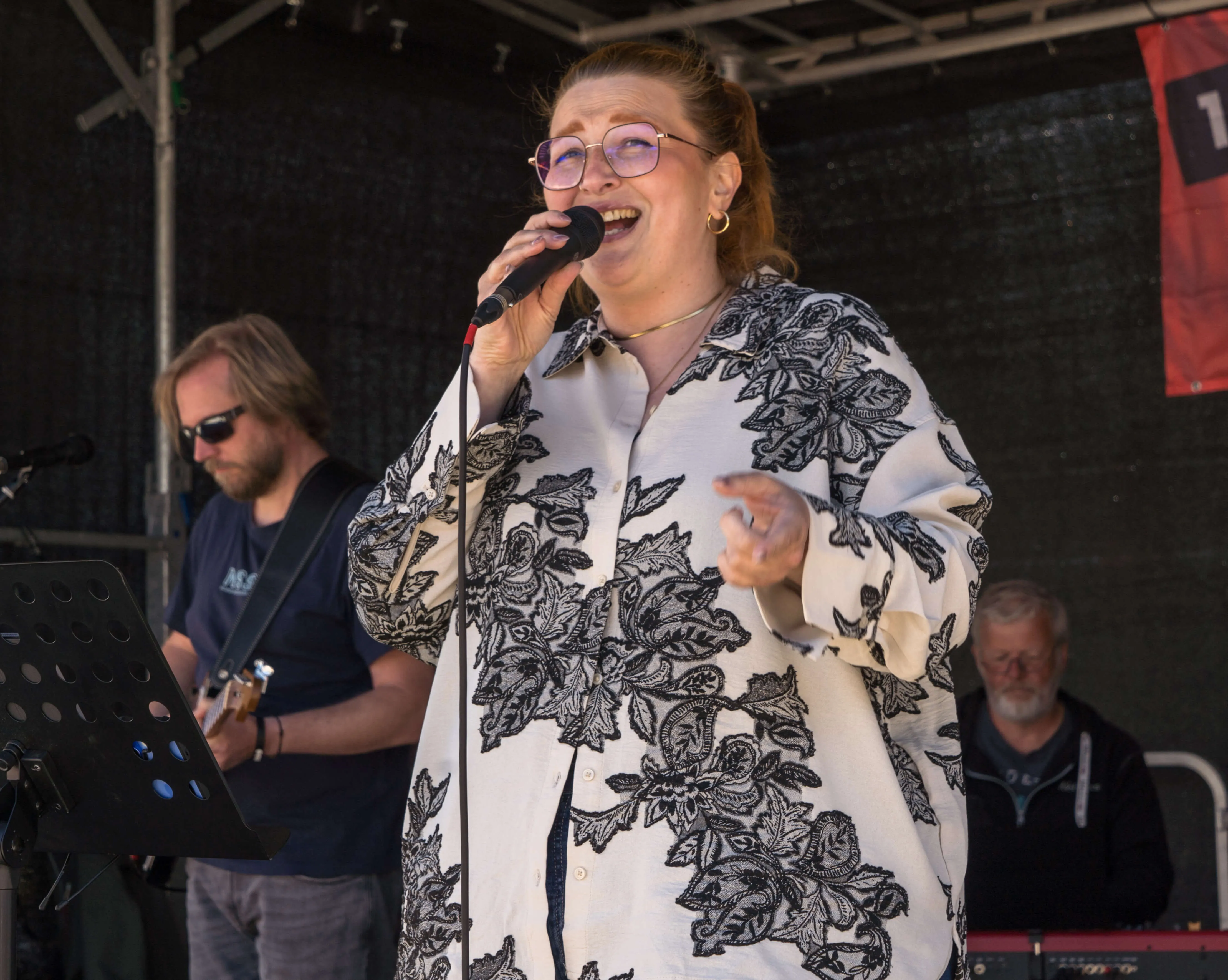 Sängerin mit rotem Haar und Brille singt in ein Mikrofon auf einer Bühne, während ein Mann mit Sonnenbrille Gitarre spielt und ein weiterer Mann im Hintergrund sitzt.