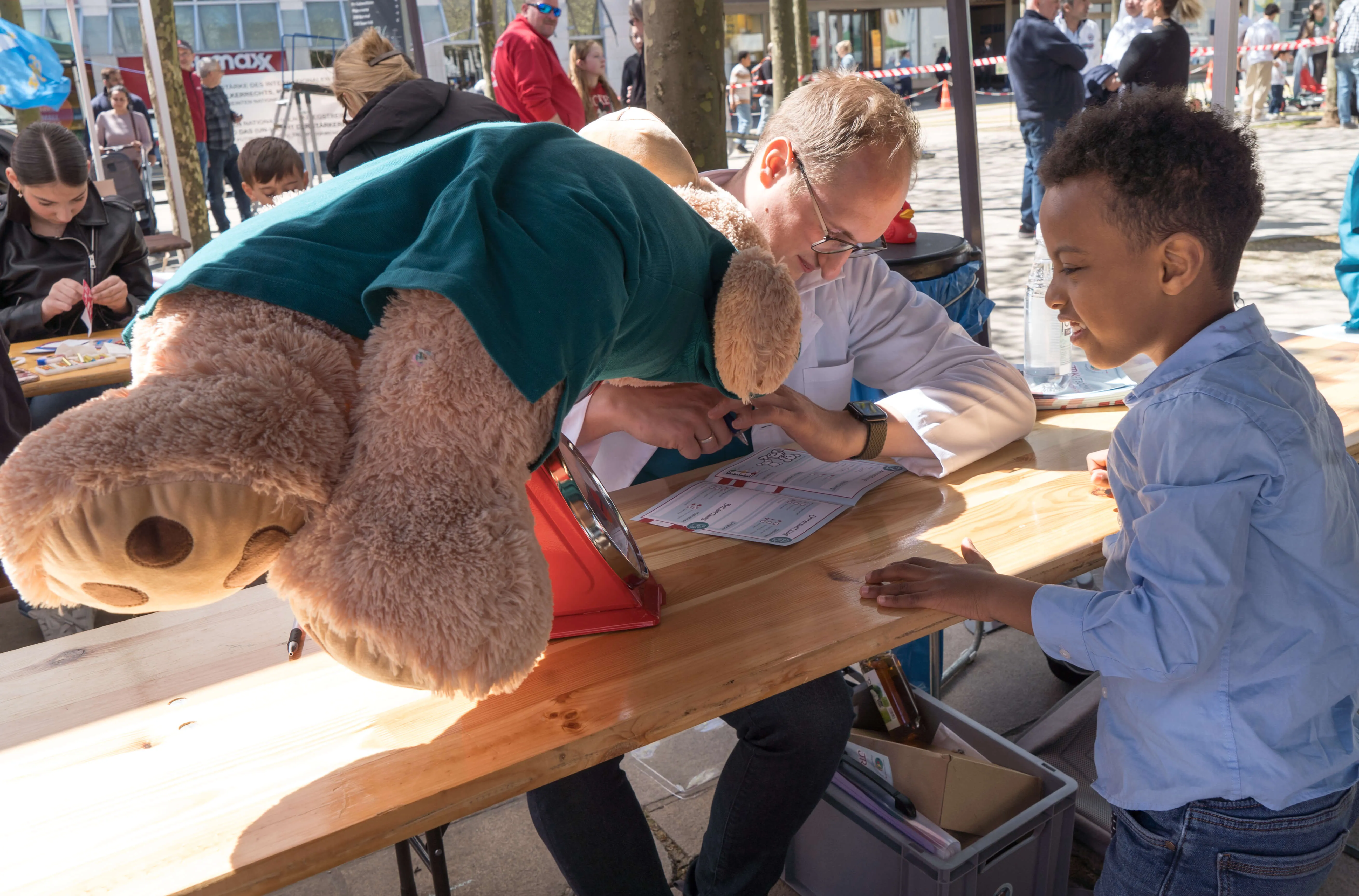 Ein Mann unterschreibt ein Tablet, während ein Kind zusieht. Im Hintergrund sitzt ein großer Teddybär am Tisch.