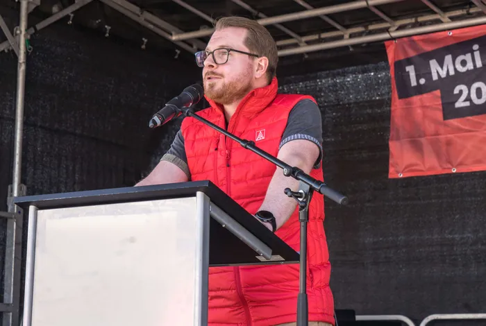 Mann mit rotem Weste hält Rede am Pult vor Publikum auf einer Mai-Kundgebung.