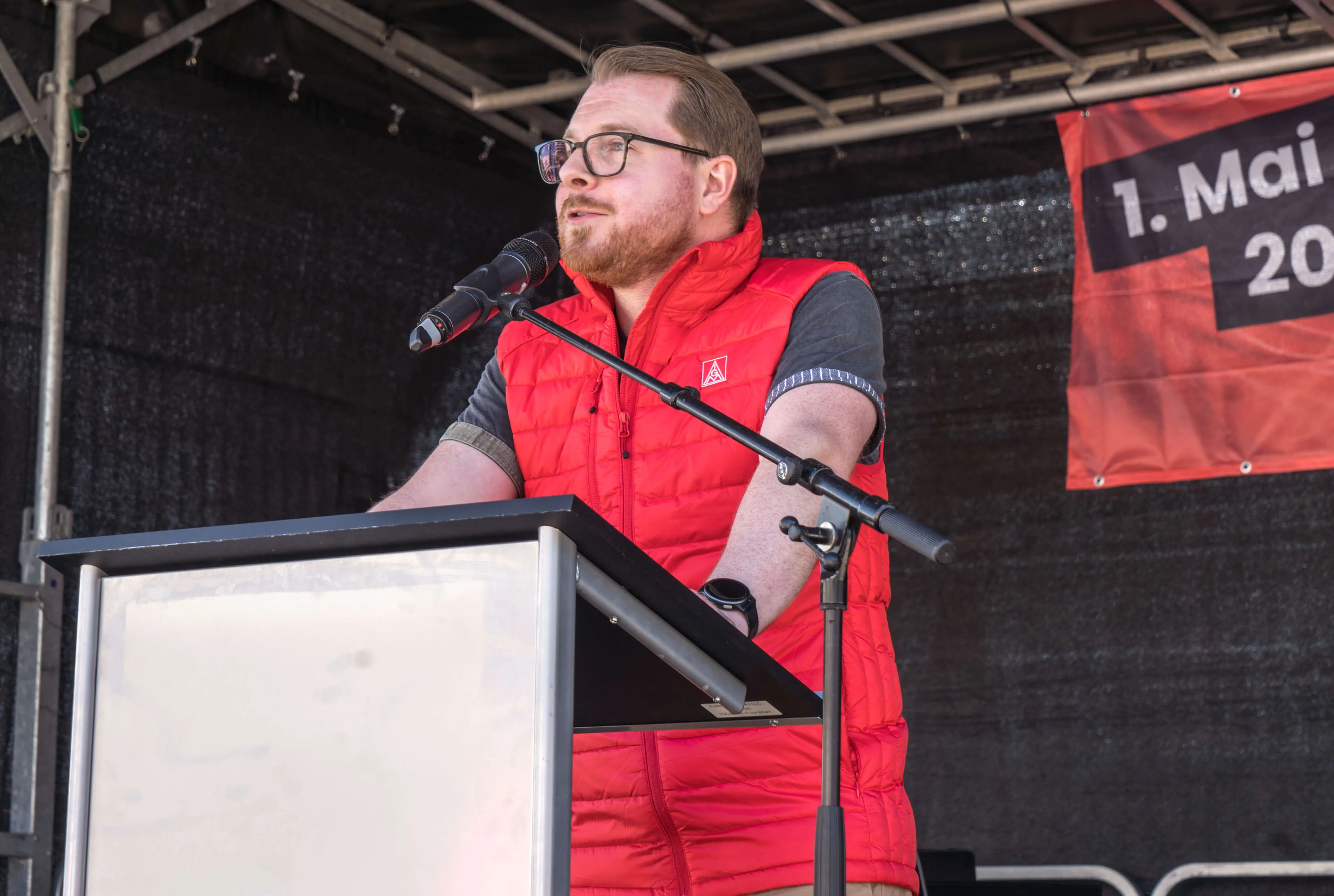 Mann mit rotem Weste hält Rede am Pult vor Publikum auf einer Mai-Kundgebung.