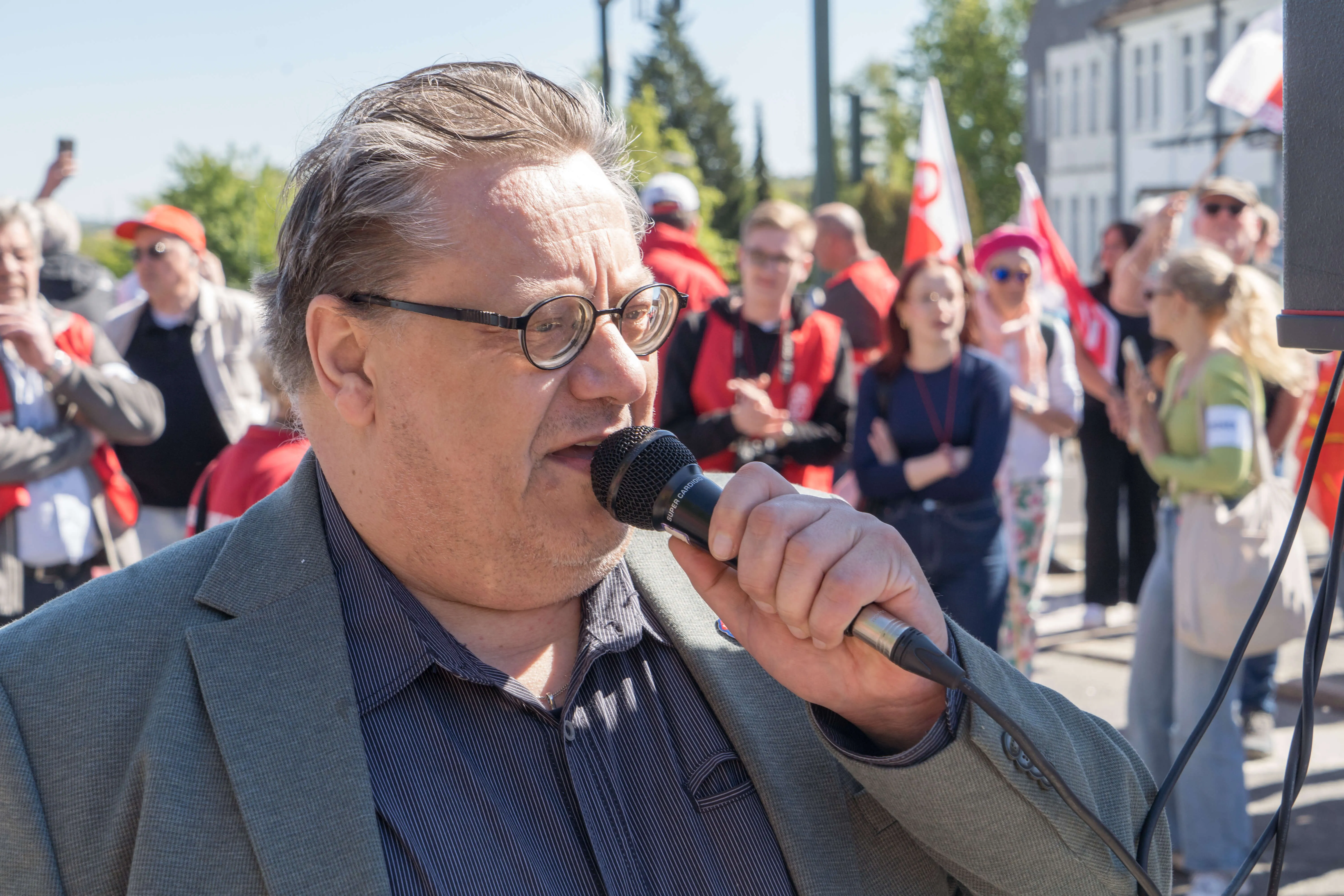 Mark Stefan Marx, Geschäftsführer des DGB Region Ruhr, spricht in einen schwarzen Handmikrofon während einer Mai-Kundgebung auf dem Lüdenscheider Rathausplatz. Im Hintergrund sind weitere Teilnehmer mit roten Fahnen und Deutschlandflaggen zu sehen.