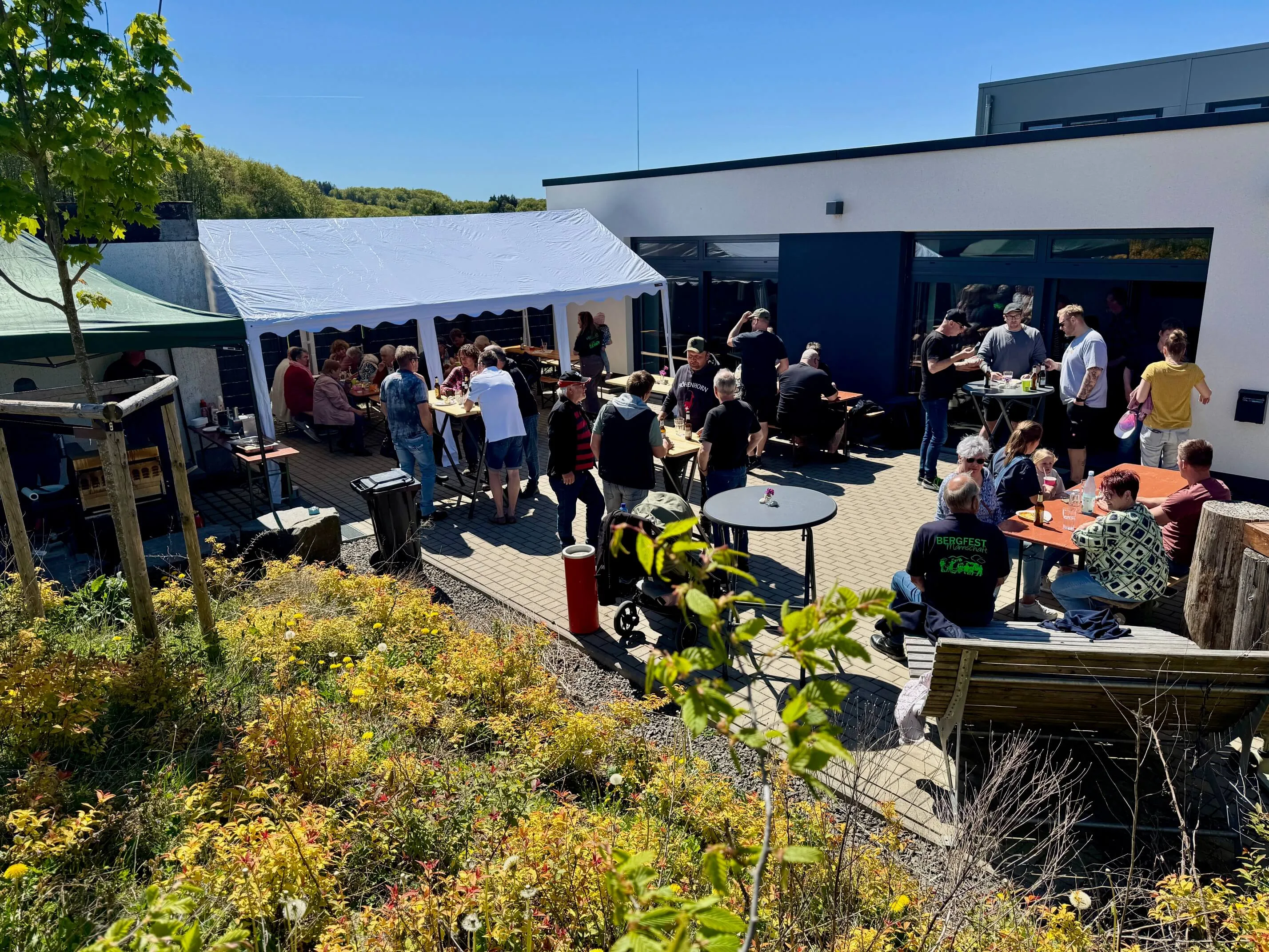 Menschen stehen und sitzen auf einem Hofgelände um einen aufgestellten Maibaum. Im Hintergrund ein Gebäude mit einer überdachten Terrasse, auf der ebenfalls Menschen sitzen.