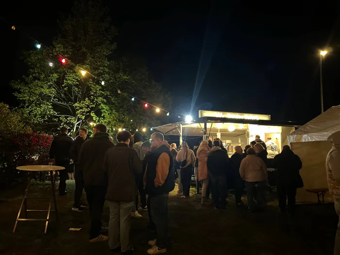 Menschen stehen vor einem beleuchteten Imbissstand auf einem nächtlichen Festgelände mit Bäumen und Lichterketten.