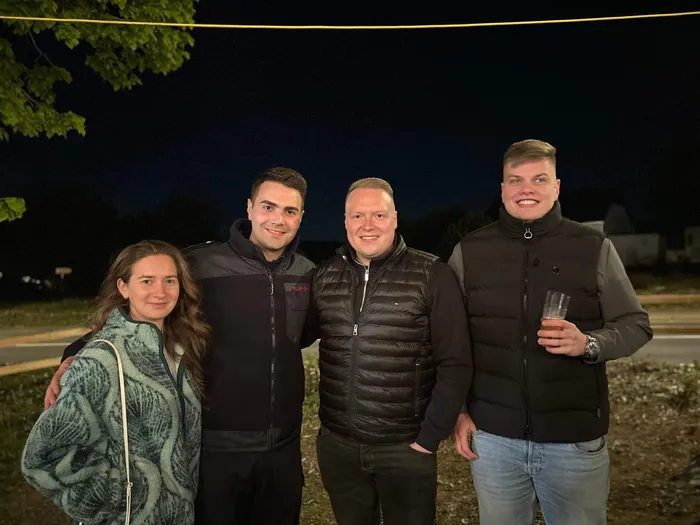 Vier junge Erwachsene stehen eng zusammen und lächeln in die Kamera. Im Hintergrund ist es dunkel, vermutlich während einer Abendveranstaltung.