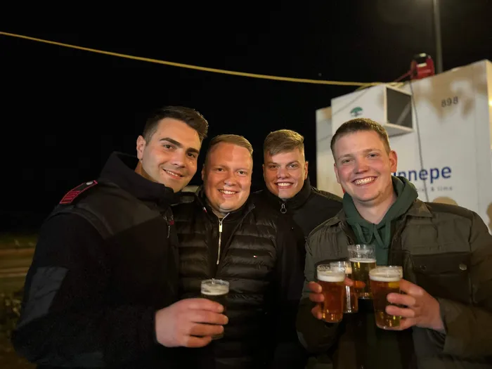 Vier junge Männer lächeln in die Kamera und halten jeweils ein Glas Bier in der Hand. Im Hintergrund ist ein beleuchteter Lastwagen mit der Aufschrift „ennepe“ zu sehen.