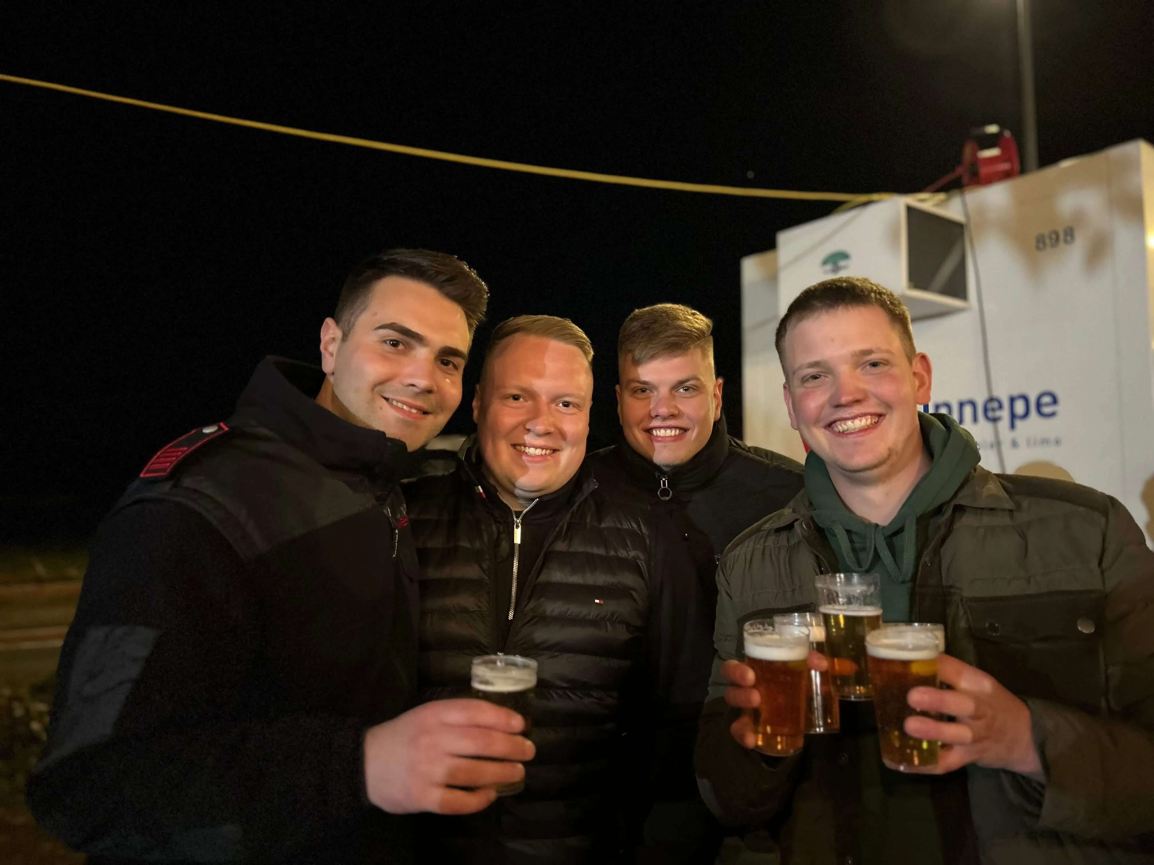 Vier junge Männer lächeln in die Kamera und halten jeweils ein Glas Bier in der Hand. Im Hintergrund ist ein beleuchteter Lastwagen mit der Aufschrift „ennepe“ zu sehen.
