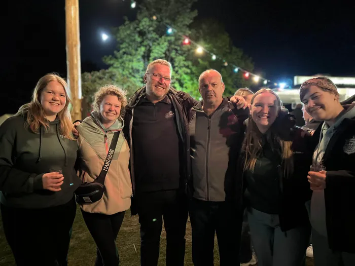 Eine Gruppe von sechs lächelnden Menschen umarmt sich vor einem verschwommenen Hintergrund mit Lichtern und Bäumen. Das Bild zeigt die gelungene Premiere des gemeinsamen Maibaumfests der Landjugend und der Kirchengemeinde Heedfeld.