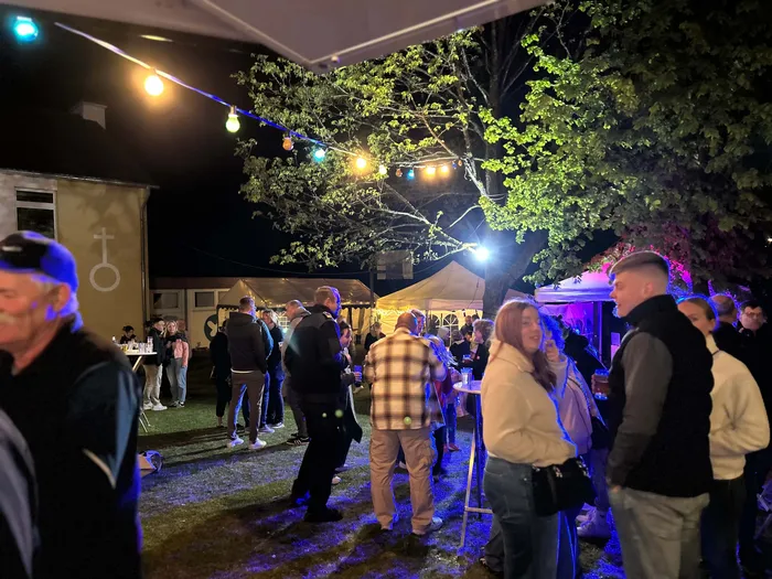 Menschen stehen und unterhalten sich im Freien vor einem Gebäude und einem Zelt, beleuchtet von Lichterketten und Lampen. Im Hintergrund ein Baum und ein dunkler Nachthimmel.