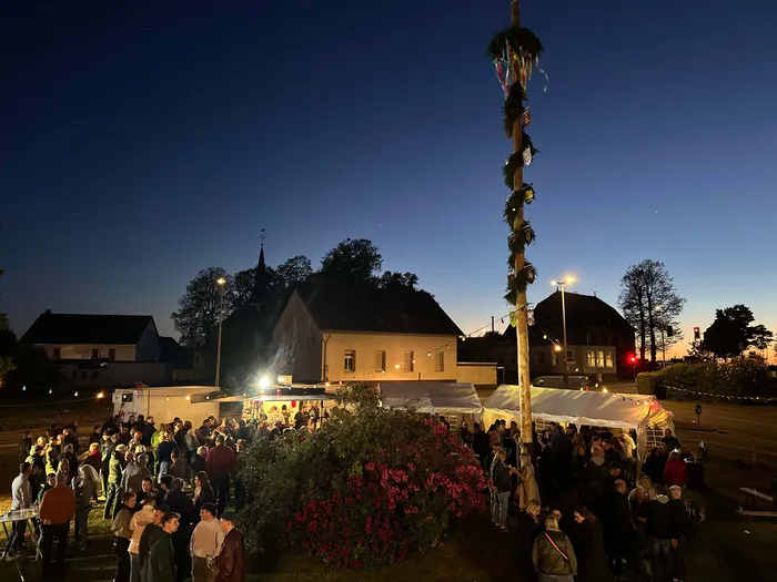 Menschengruppe vor einem Maibaum und einem festlich beleuchteten Imbissstand vor einer Kirche bei Dämmerung.
