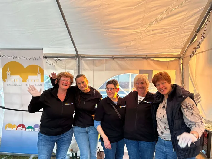 Fünf lächelnde Frauen stehen eng zusammen unter einem Zelt. Im Hintergrund ist ein Banner mit den Logos der Kirchengemeinde Hülscheid und Heedfeld zu sehen.