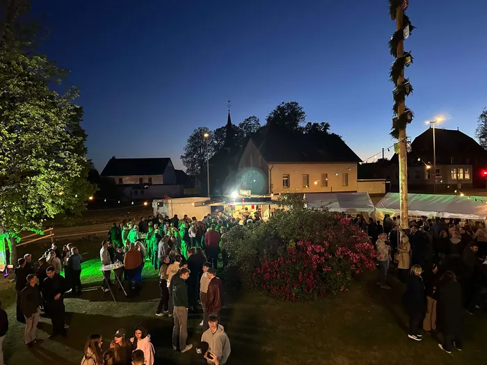 Menschen stehen und unterhalten sich auf einer Wiese vor einem hell erleuchteten Gebäude mit einem Kirchturm im Hintergrund während des Maibaumfestes.