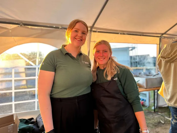 Zwei junge Frauen lächeln in grünen Poloshirts und einer Schürze unter einem Zelt. Im Hintergrund sind weitere Personen und ein Tisch zu sehen.