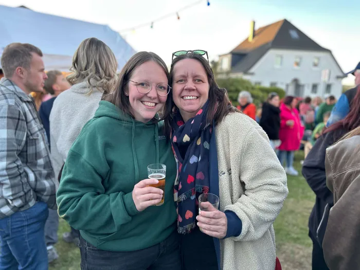 Zwei lächelnde Frauen umarmen sich auf dem Maibaumfest in Heedfeld, beide halten Gläser in der Hand. Im Hintergrund sind weitere Menschen und Häuser zu sehen.