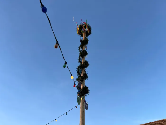 Hoher, geschmückter Maibaum mit bunten Bändern und Lichterketten vor blauem Himmel.