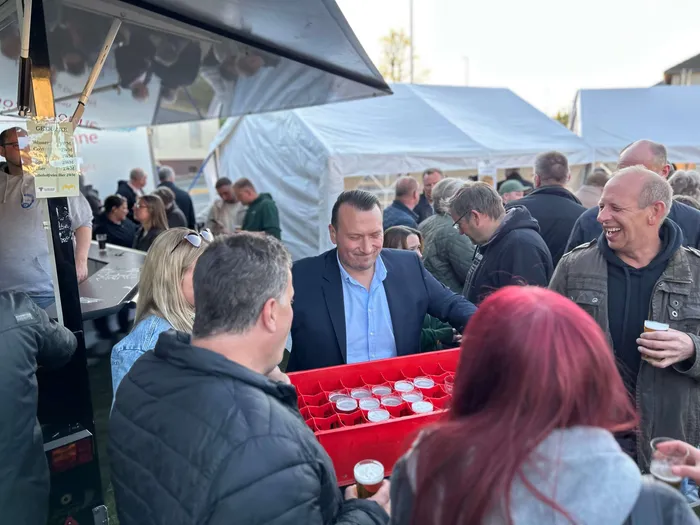 Menschen stehen um einen Tresen mit Getränken und kleinen Snacks auf einem Freiluftfest, vermutlich einem Maibaumfest. Im Hintergrund sind weitere Besucher und Zelte zu sehen.