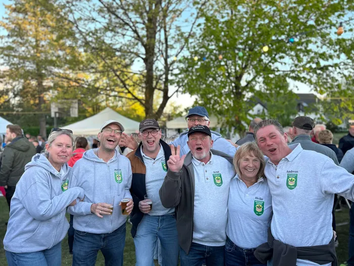 Eine Gruppe von Menschen in Freizeitkleidung steht lächelnd vor einem Maibaum, einige halten Bierkrüge in der Hand.