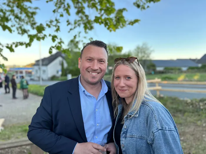 Ein Mann und eine Frau lächeln in die Kamera. Im Hintergrund sind Häuser und grüne Flächen zu sehen.