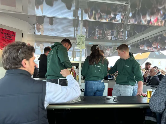 Mehrere junge Männer in grünen Hoodies bedienen einen Getränkestand auf dem Maibaumfest in Heedfeld, während ein Mann im Vordergrund ein Bierglas entgegennimmt.