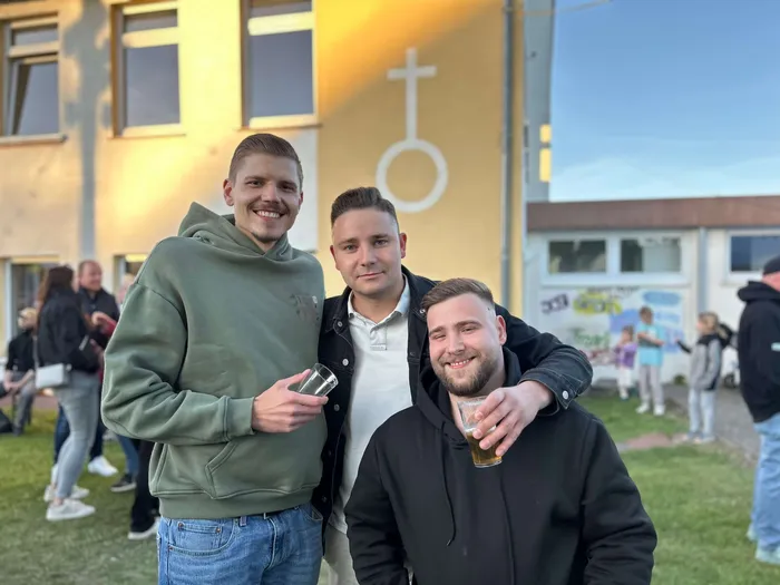 Drei junge Männer umarmen sich vor einem hellgelben Gebäude mit einem stilisierten Kreuz. Im Hintergrund sind weitere Menschen und ein bunt bemaltes Fenster zu sehen.