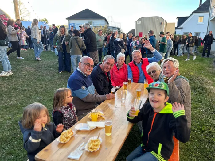 Menschen sitzen und stehen bei einem Maibaumfest zusammen, einige essen Pommes und trinken Bier an einem Holztisch. Im Hintergrund eine Menschenmenge und Gebäude.