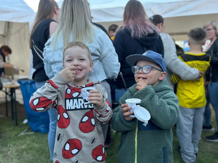 Zwei lächelnde Jungen essen Joghurt auf einem Volksfest, einer trägt ein Spiderman-Shirt und der andere eine Baseballkappe.