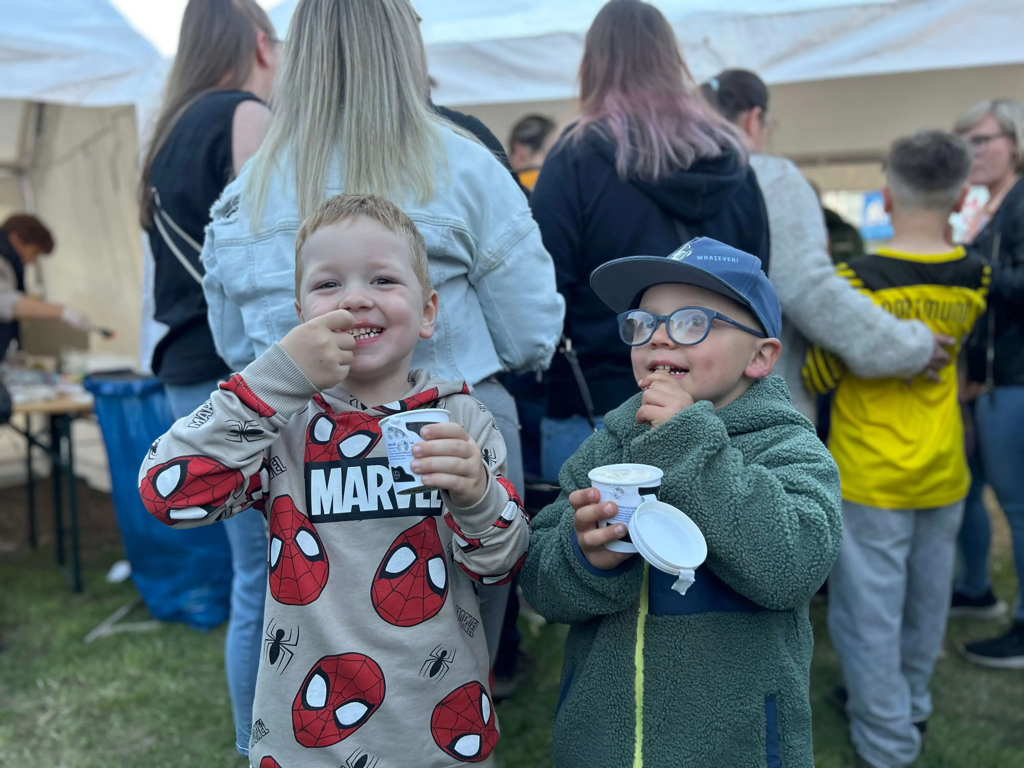 Zwei lächelnde Jungen essen Joghurt auf einem Volksfest, einer trägt ein Spiderman-Shirt und der andere eine Baseballkappe.